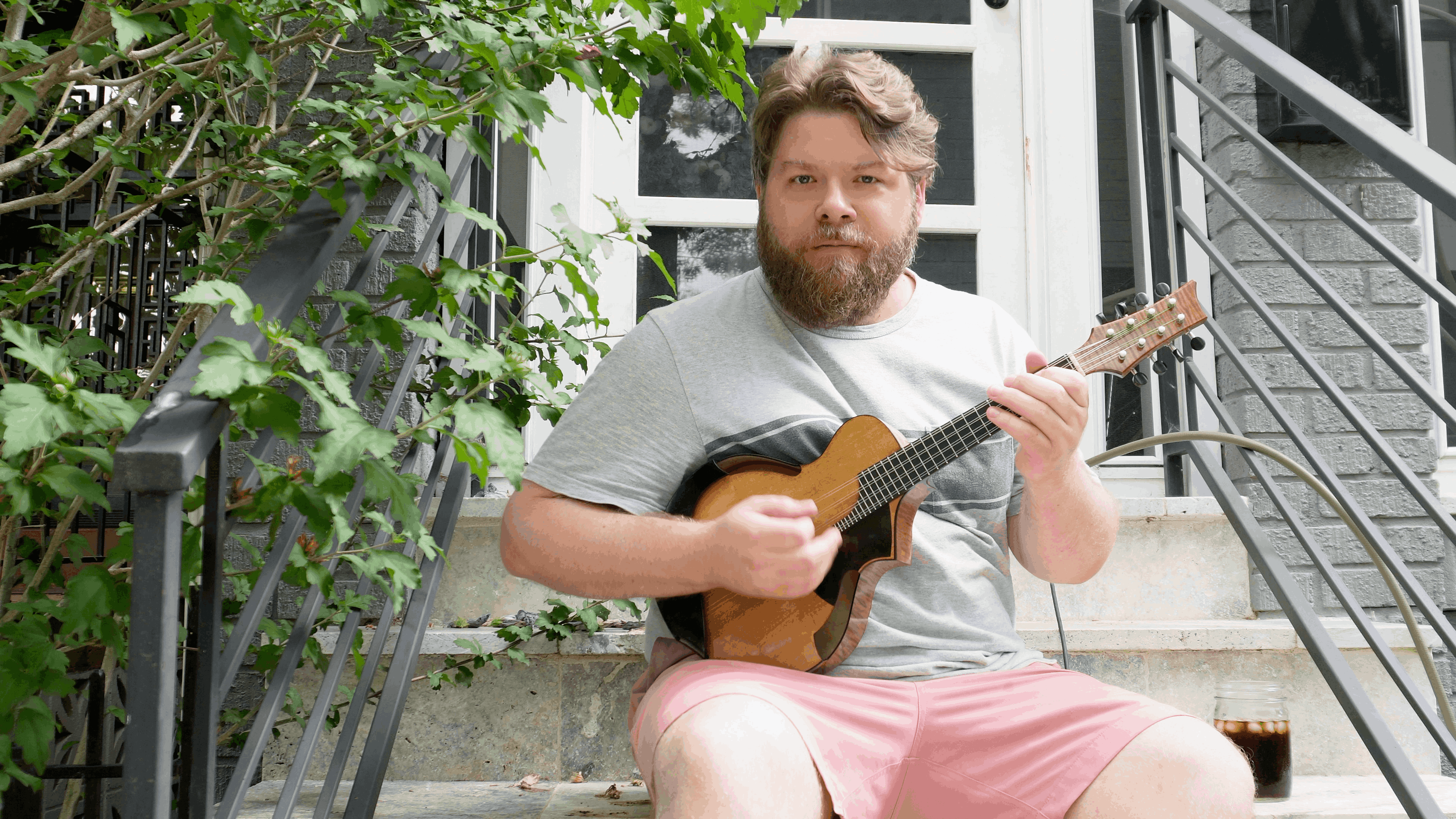 Josh playing mandolin on his porch stairs next to ice coffee with a green tree with like 2 small pink flowers